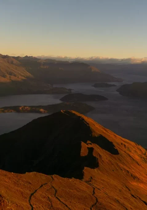 A wide view from Roys Peak at sunrise, showing the winding hiking trail along the golden-lit ridgeline with Lake Wānaka and its islands below. The surrounding mountains are partially cast in shadow with soft morning light hitting the peaks.