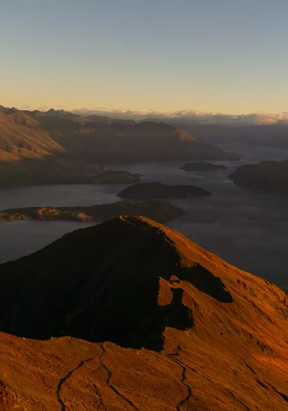 A wide view from Roys Peak at sunrise, showing the winding hiking trail along the golden-lit ridgeline with Lake Wānaka and its islands below. The surrounding mountains are partially cast in shadow with soft morning light hitting the peaks.