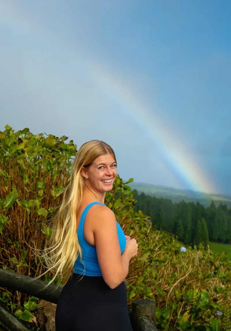 A woman standing by a wooden railing with a rainbow visible over a lush green valley in the Azores.