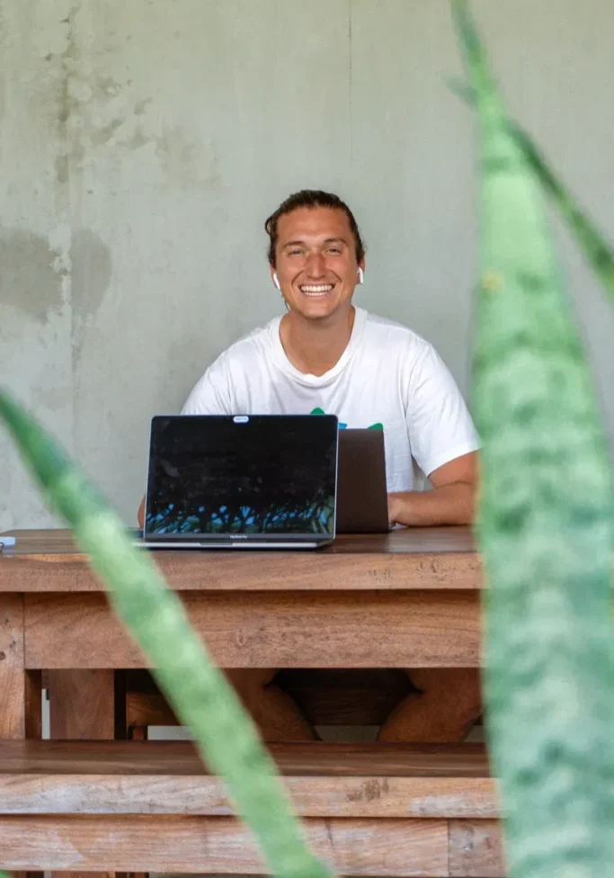 Smiling digital nomad working remotely with a laptop at a wooden table in Sri Lanka, framed by green tropical plants in the foreground.