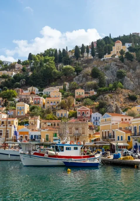 Colourful buildings on the hillside in Symi, Greece, with fishing boats docked along the vibrant waterfront under a bright blue sky. The pastel homes, set against rocky terrain and cypress trees, create a picturesque Mediterranean harbour scene.