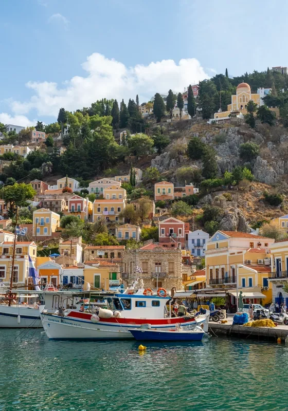 Colourful buildings on the hillside in Symi, Greece, with fishing boats docked along the vibrant waterfront under a bright blue sky. The pastel homes, set against rocky terrain and cypress trees, create a picturesque Mediterranean harbour scene.