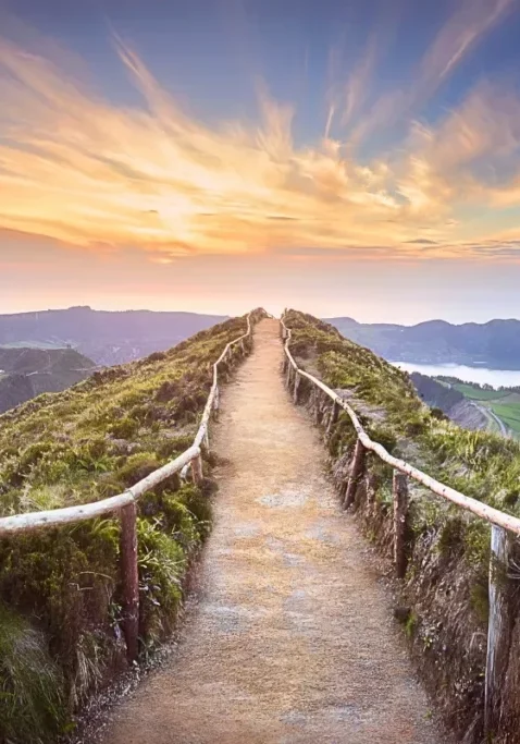 A hiking trail with rustic wooden railings leads along a ridgeline at sunset, overlooking the Sete Cidades crater lakes on São Miguel Island in the Azores. The glowing sky and sweeping coastal views highlight one of the most iconic hiking spots in the archipelago.