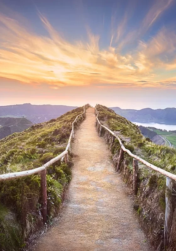 A hiking trail with rustic wooden railings leads along a ridgeline at sunset, overlooking the Sete Cidades crater lakes on São Miguel Island in the Azores. The glowing sky and sweeping coastal views highlight one of the most iconic hiking spots in the archipelago.