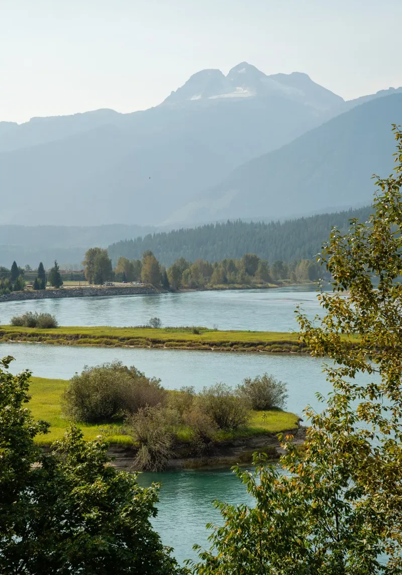 A beautiful view of the Columbia River winding through lush, tree-lined islands with Mt Begbie rising in the background, its glacier-topped summit softened by fall haze. Early autumn colors frame the scene, highlighting Revelstoke’s serene natural beauty.