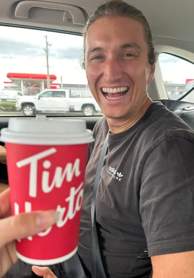 A man sitting in the seat of a car, smiling widely. In the foreground, a hand holds a red Tim Hortons coffee cup toward the camera. Through the window, a gas station and parked vehicles are visible outside.