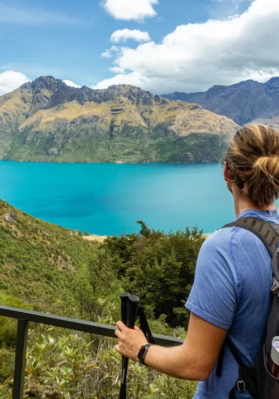 A hiker with a backpack and walking pole stands at a lookout point on the Wye Creek Track, looking out over the vivid turquoise waters of Lake Wakatipu. Towering mountains with rugged peaks rise in the background under a partly cloudy sky, surrounded by lush greenery.