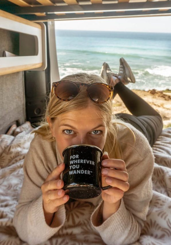A girl laying in her camper van while enjoying a cup of coffee.