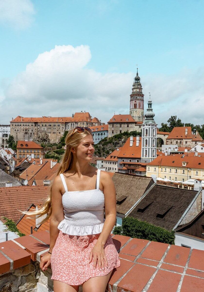 A woman in a white top and pink floral skirt stands in front of the orange rooftops and historic buildings of Cesky Krumlov, with the castle tower rising in the background under a blue sky.